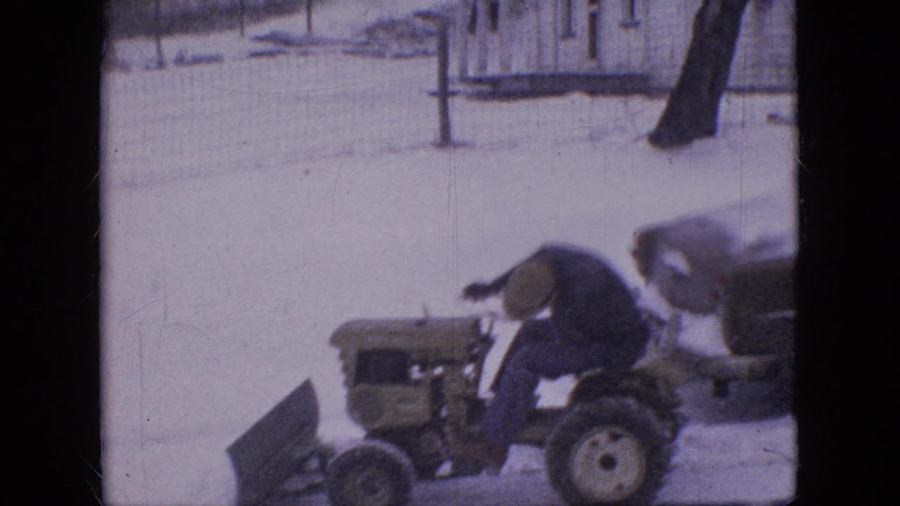 In this 1964 Philadelphia home-movie frame, a man in winter clothing operates a small tractor with a front snowplow blade through snow beside a wire fence.
