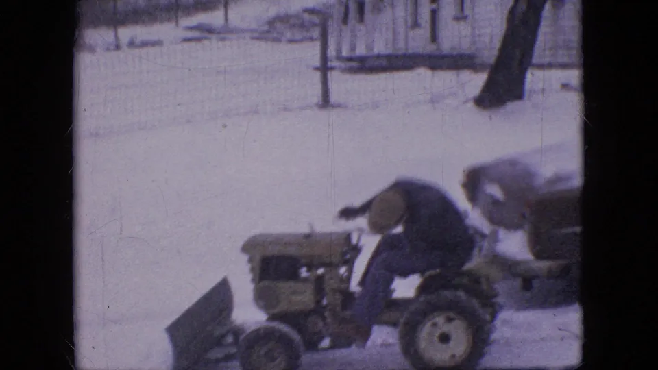 In this 1964 Philadelphia home-movie frame, a man in winter clothing operates a small tractor with a front snowplow blade through snow beside a wire fence.
