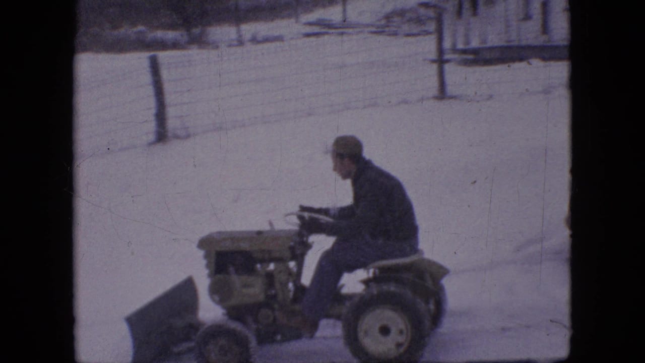 A man in a cap and dark coat sits on a compact tractor with a front plow blade in the snowy Philadelphia residential setting described by the clip metadata.