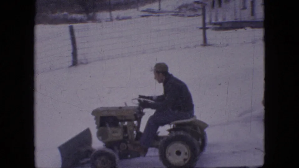 A man in a cap and dark coat sits on a compact tractor with a front plow blade in the snowy Philadelphia residential setting described by the clip metadata.
