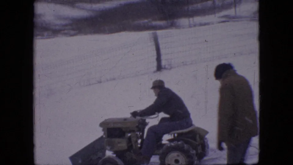 The snowplow tractor moves through the snowy residential area as another bundled adult stands nearby, captured in soft 8mm home-movie focus.
