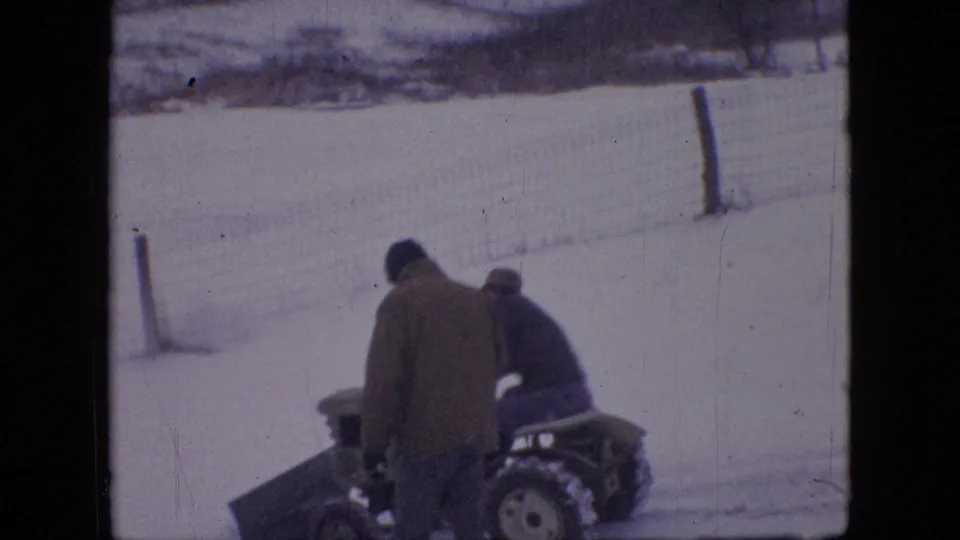 A bundled adult stands beside the seated tractor driver near the front plow blade, with snow, fencing, and the soft color of 8mm film visible around them.
