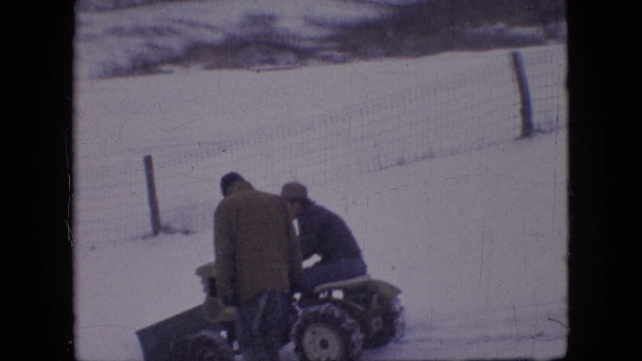 In 1964 Philadelphia-area 8mm home-movie footage, two adult men in winter coats stand near a compact tractor fitted with a front snow plow on snow-covered ground.