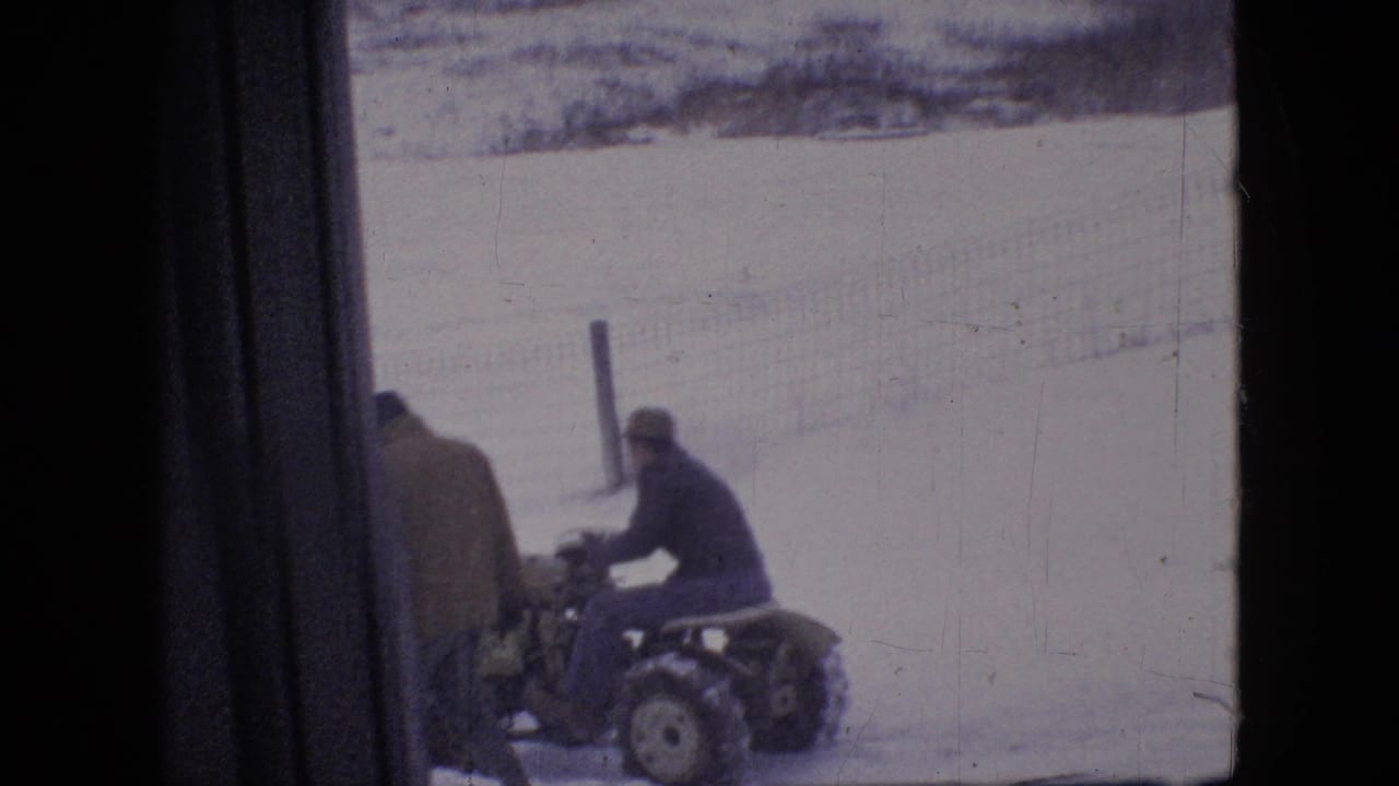 A man appears seated on a compact snow plow tractor while another man in a heavy coat stands nearby in a snowy Philadelphia-area outdoor setting from 1964 metadata.