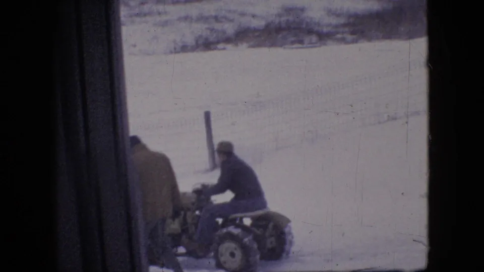 A man appears seated on a compact snow plow tractor while another man in a heavy coat stands nearby in a snowy Philadelphia-area outdoor setting from 1964 metadata.