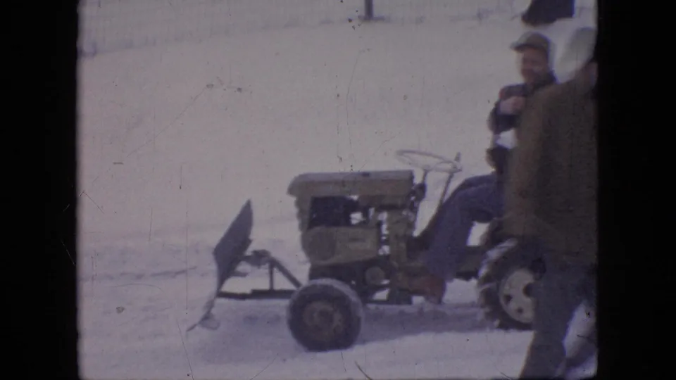 The frame gives a closer 8mm view of the compact tractor and angled snow plow blade, with adult men partly visible at the edge of the snowy scene.