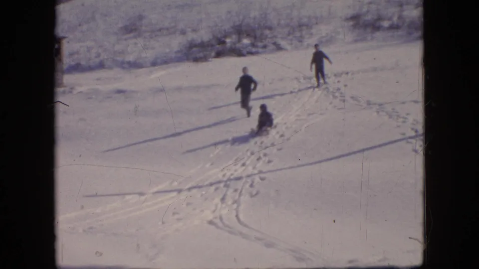 A wider 1964 home-movie frame shows people moving across a snow-covered area with footprints or tracks visible, likely near the same winter plowing scene.