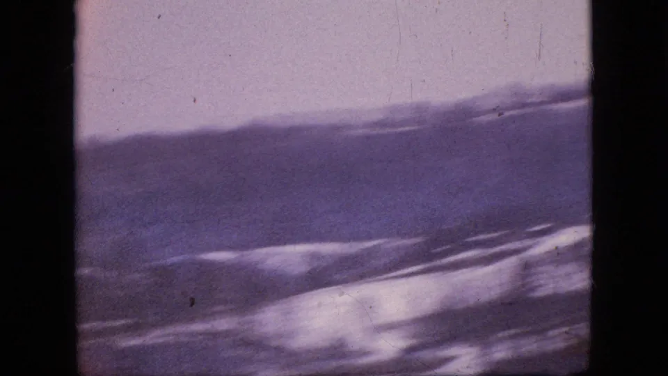 A grainy 8mm frame of rolling snow-covered hills beneath a pale sky, with muted purple tones and visible scratches or dust in the film scan.