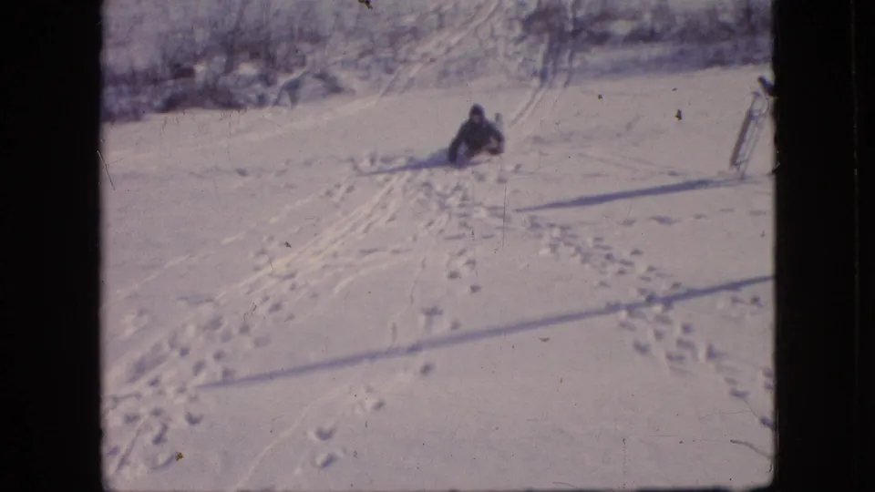 A bundled figure sits or comes to rest on the snowy hill in the 1964 Philadelphia footage, with sled marks, footprints, and part of a sled visible near the frame edge.