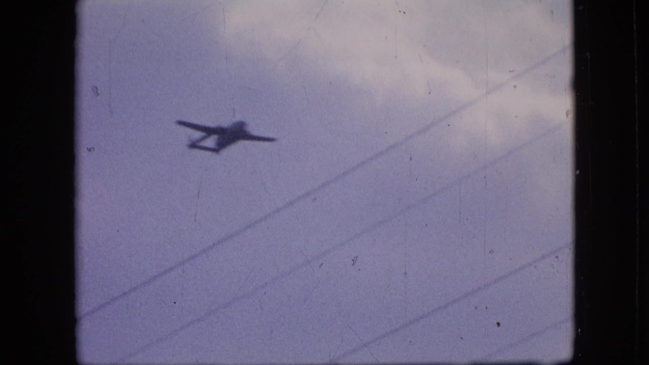 A 1964 Philadelphia 8mm frame shows a small dark twin-boom airplane silhouette moving across a pale overcast sky with diagonal utility wires in the foreground.