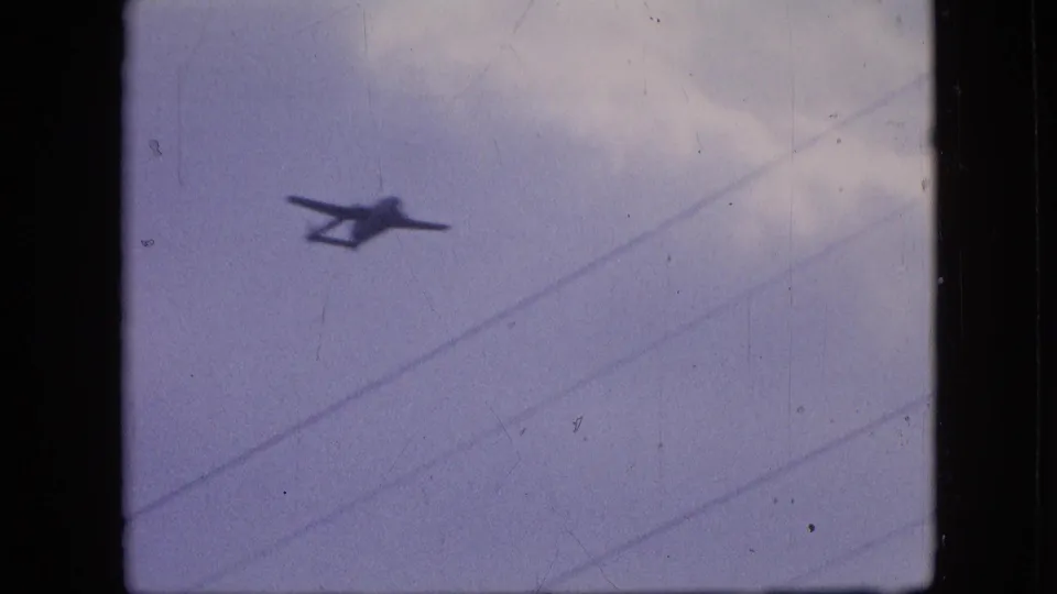 A 1964 Philadelphia 8mm frame shows a small dark twin-boom airplane silhouette moving across a pale overcast sky with diagonal utility wires in the foreground.