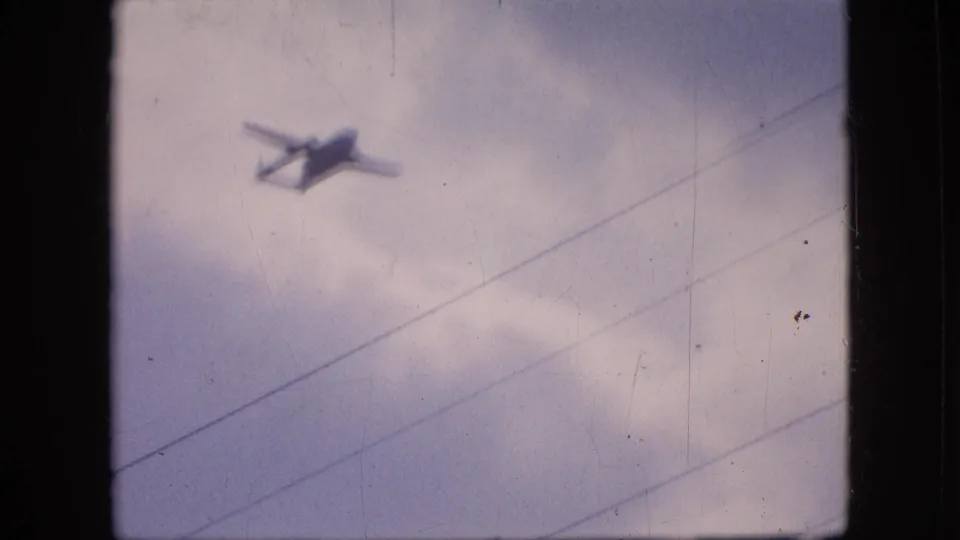The aircraft appears near the upper part of the 1964 Philadelphia frame, silhouetted against pale clouds while diagonal wires cut across the sky.