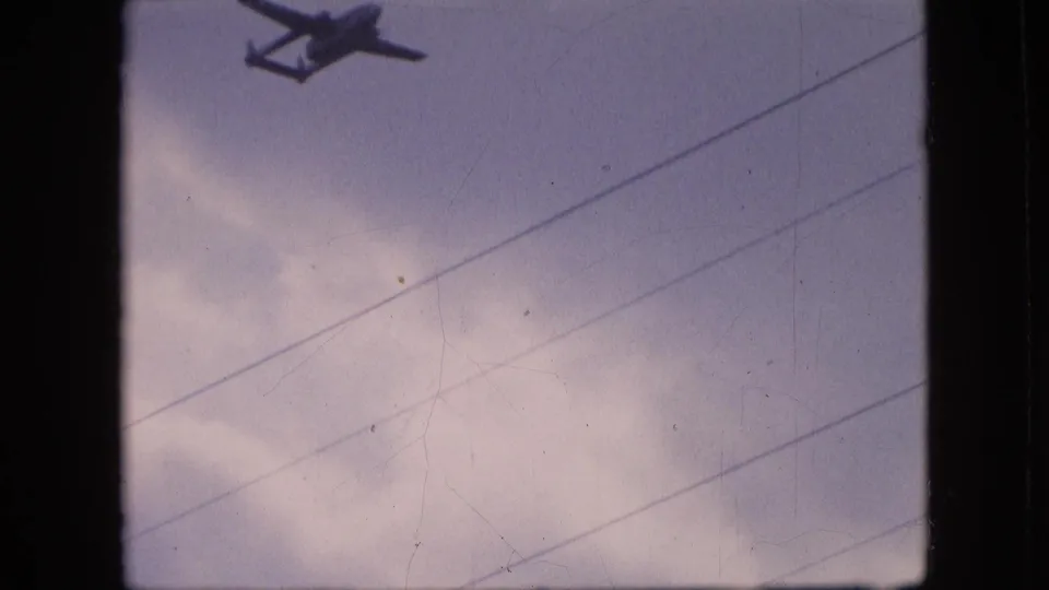A dark aircraft silhouette sits near the upper-left edge of the 8mm frame, with overcast clouds, diagonal utility wires, and visible film wear across the view.