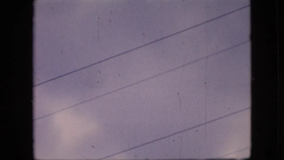 1964 Philadelphia home-movie frame showing a cloudy pastel sky crossed by diagonal overhead electric wires and framed by dark 8mm gate edges.