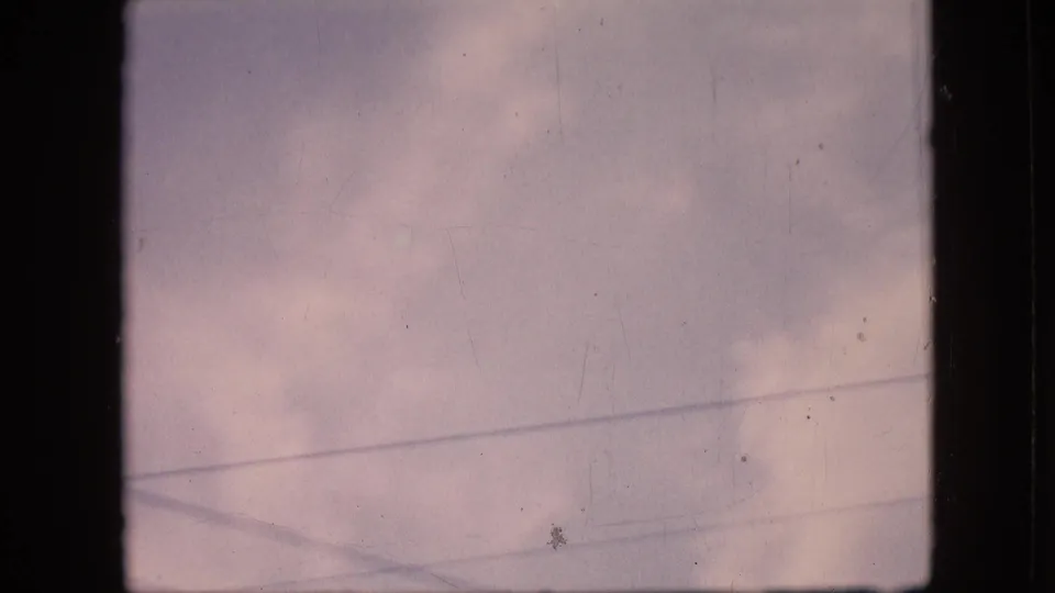 Philadelphia home-movie sky frame from 1964 showing pale clouds, crossing electric wires, and visible scratches and dust in the film surface.