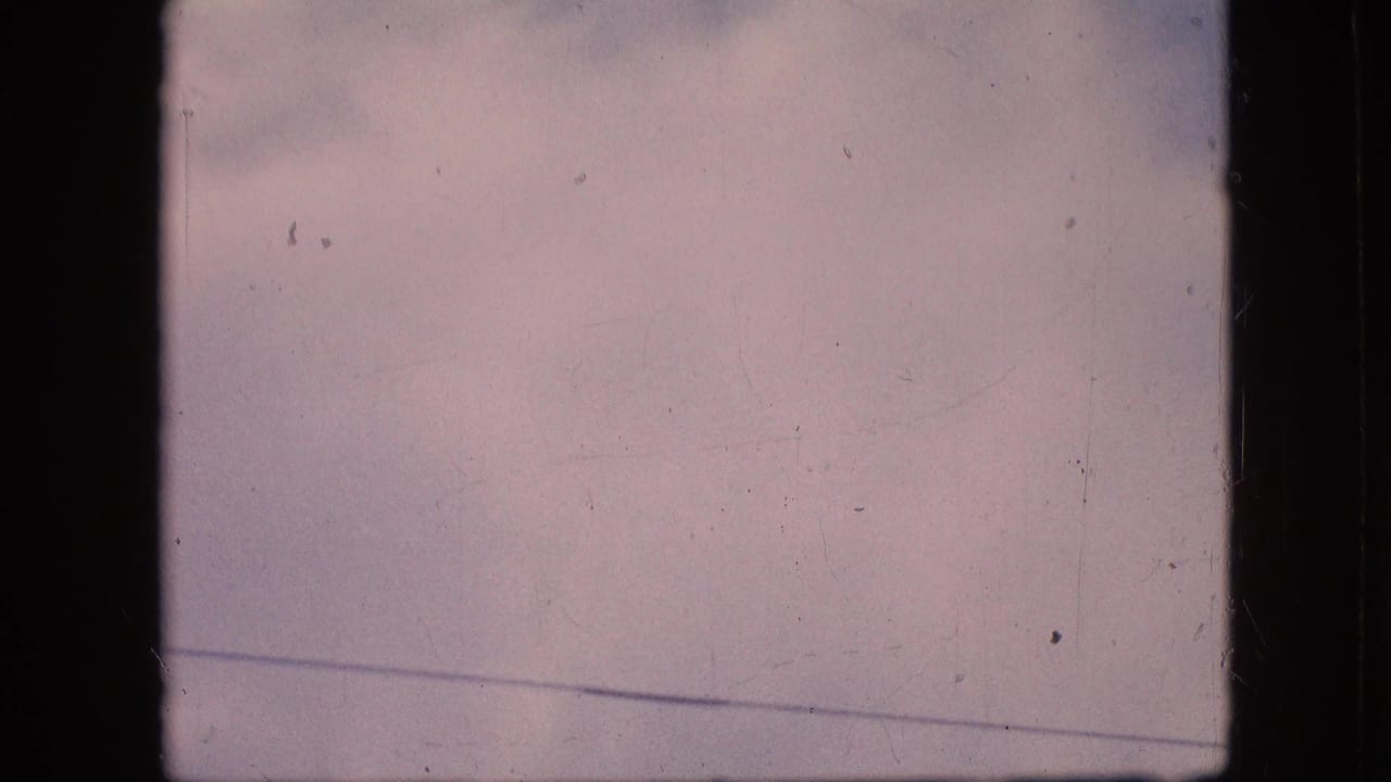1964 Philadelphia home-movie frame showing pale pink-blue sky, faint cloud cover, visible 8mm borders, dust, and scratches before the aircraft enters view.