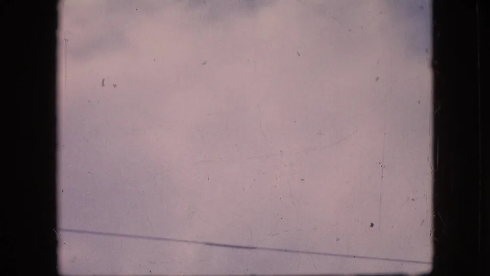 1964 Philadelphia home-movie frame showing pale pink-blue sky, faint cloud cover, visible 8mm borders, dust, and scratches before the aircraft enters view.