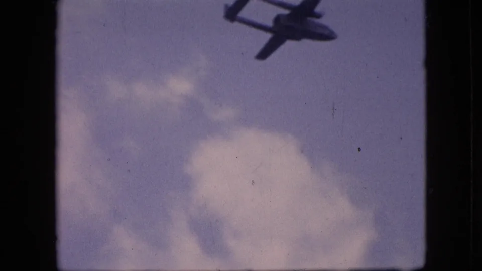 1964 Philadelphia home-movie frame with a military-style aircraft silhouette partly cropped near the top edge against a cloudy blue sky.