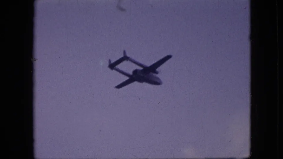 1964 Philadelphia home-movie frame showing a dark twin-boom aircraft silhouette crossing a faded blue sky with visible 8mm grain and dust.