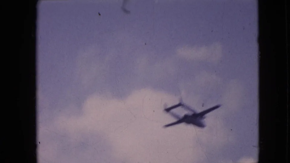 1964 Philadelphia home-movie frame with a small dark aircraft silhouette moving across a pale blue sky beneath soft clouds and visible film wear.