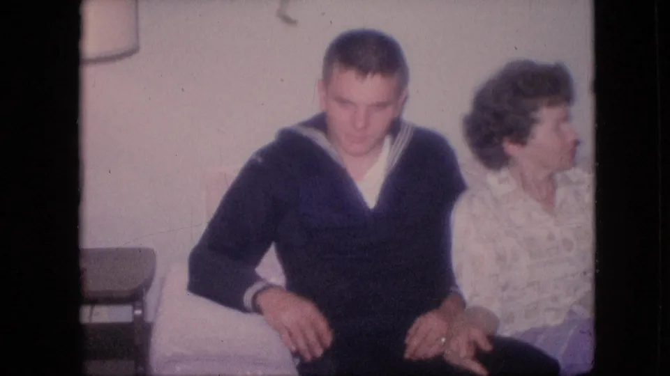 Young Navy sailor in a dark uniform with white trim sits beside a woman in a light patterned dress inside a domestic room, consistent with the supplied 1964 Philadelphia metadata.