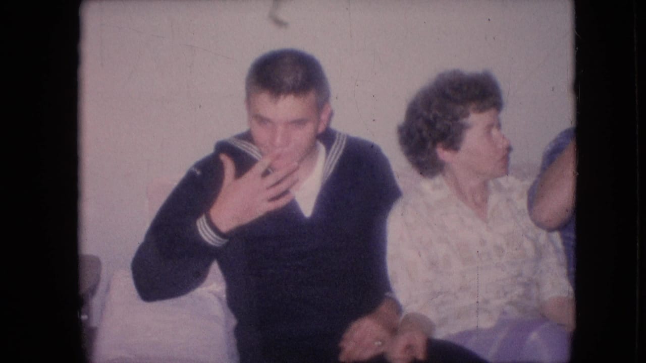 1964 Philadelphia home-movie frame showing adults seated closely on a couch, including a young man in a navy sailor-style sweater covering his mouth beside a woman in a light patterned blouse.