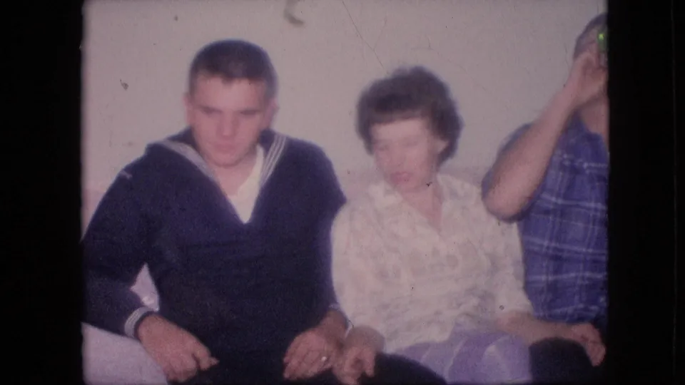 1964 Philadelphia home-movie frame showing a seated group on a couch, with a young man in a dark sailor-style sweater, a woman in a light blouse, and a cropped person at the right edge.