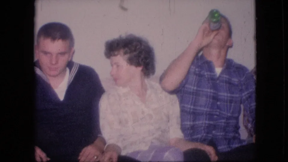 1964 Philadelphia home-movie frame showing adults on a couch as a man in a blue plaid shirt drinks from a green bottle beside a woman in a patterned blouse.