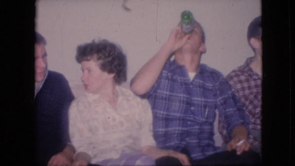 1964 Philadelphia home-movie frame showing a man in a blue plaid shirt drinking from a green bottle, with a woman and other seated adults partially visible in a private interior.