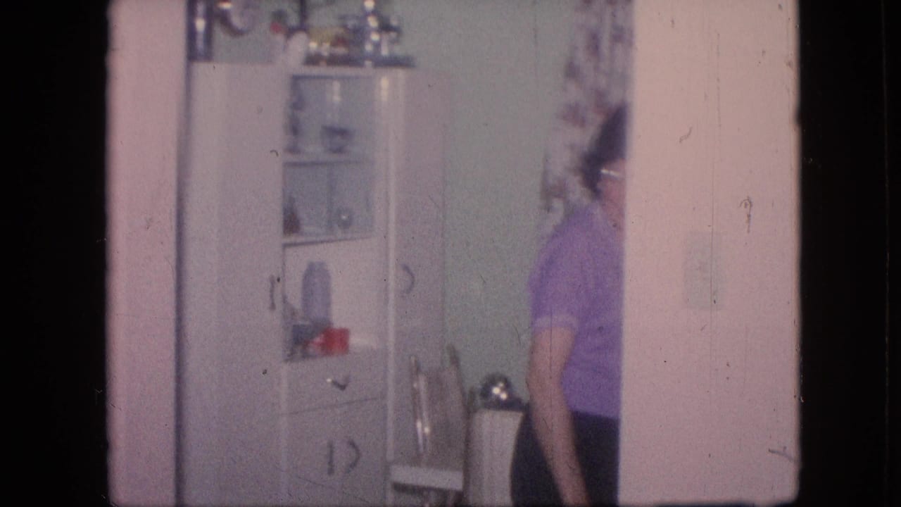 1964 Philadelphia home-movie frame showing a woman in a purple top moving near a white glass-front kitchen cabinet with dishes inside.