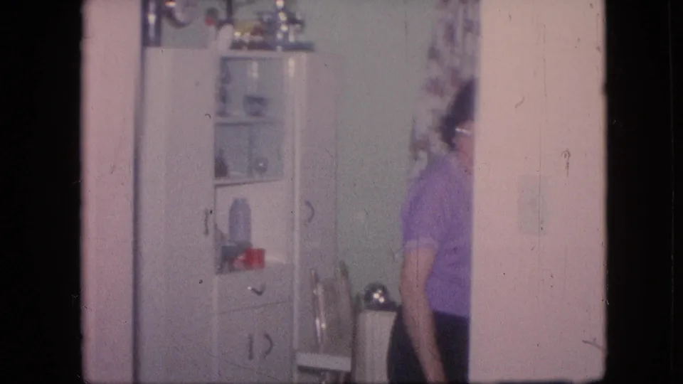 1964 Philadelphia home-movie frame showing a woman in a purple top moving near a white glass-front kitchen cabinet with dishes inside.