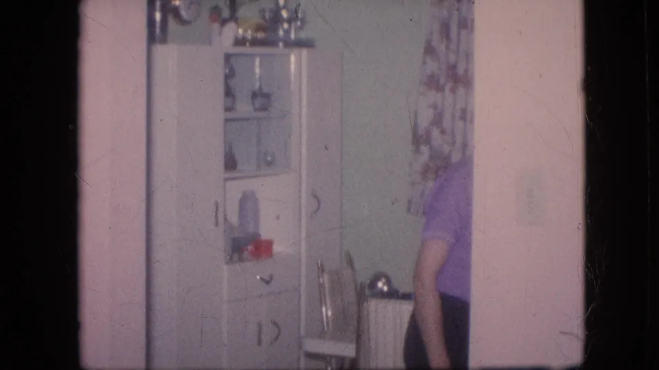 A woman in a purple top stands partly cropped at the edge of a domestic kitchen area with a white cabinet, shelves, and patterned curtain visible.