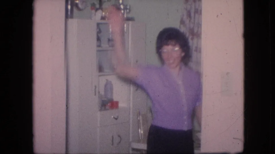 1964 Philadelphia home-movie frame showing a woman in a purple top facing the camera with one arm raised in front of a white kitchen cabinet.