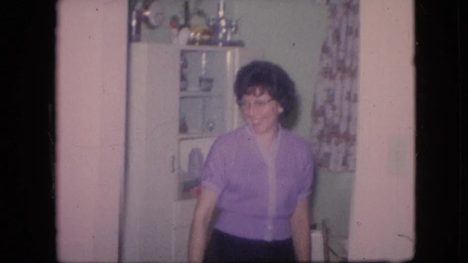A woman in a purple top stands near a white glass-front cabinet in a private kitchen interior, with soft 8mm focus and visible film borders.