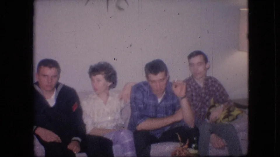 1964 Philadelphia home-movie frame showing three men and one woman seated on a light couch, with the group facing forward and toward one another in a private living room.