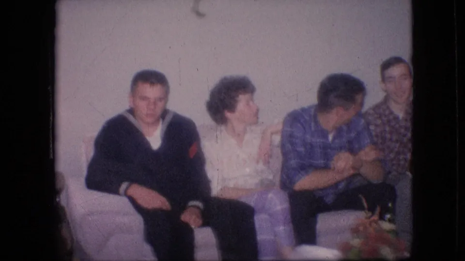 1964 Philadelphia home-movie frame showing a seated group in conversation, including a woman turned toward a man in a blue plaid shirt while another man looks toward the camera.