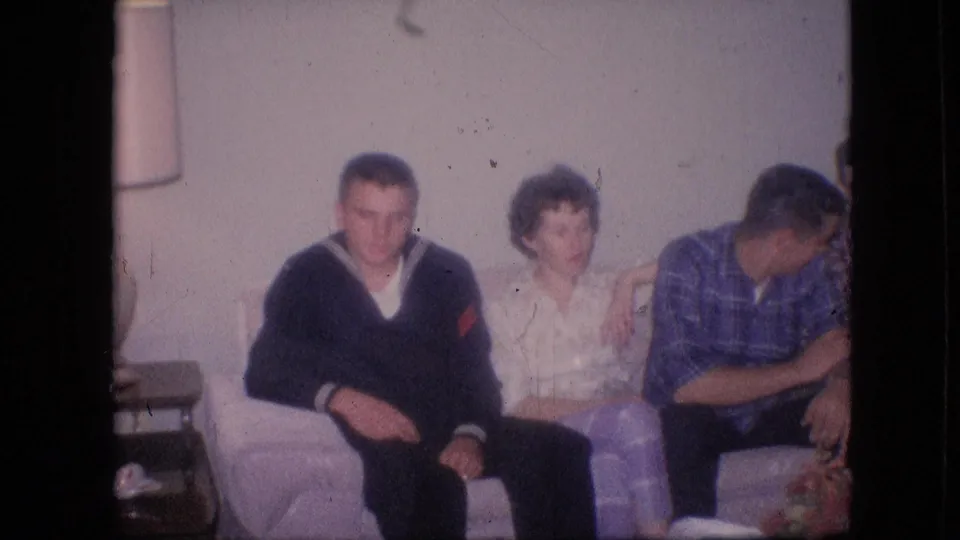 1964 Philadelphia home-movie frame showing adults seated on a light couch, with a table lamp and side table visible at the left edge of the living room.