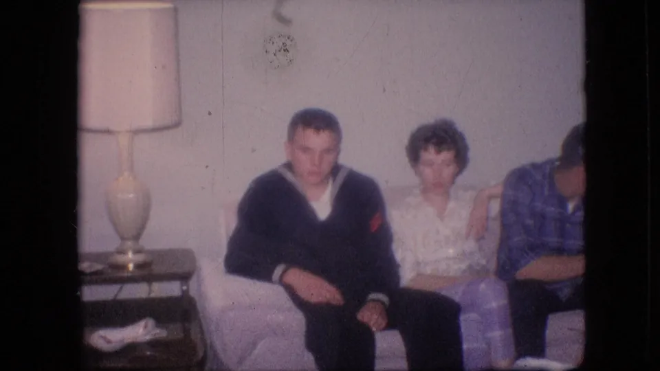 1964 Philadelphia home-movie frame showing two adults centered on a light couch, a third partly visible at right, and a table lamp beside the seating area.
