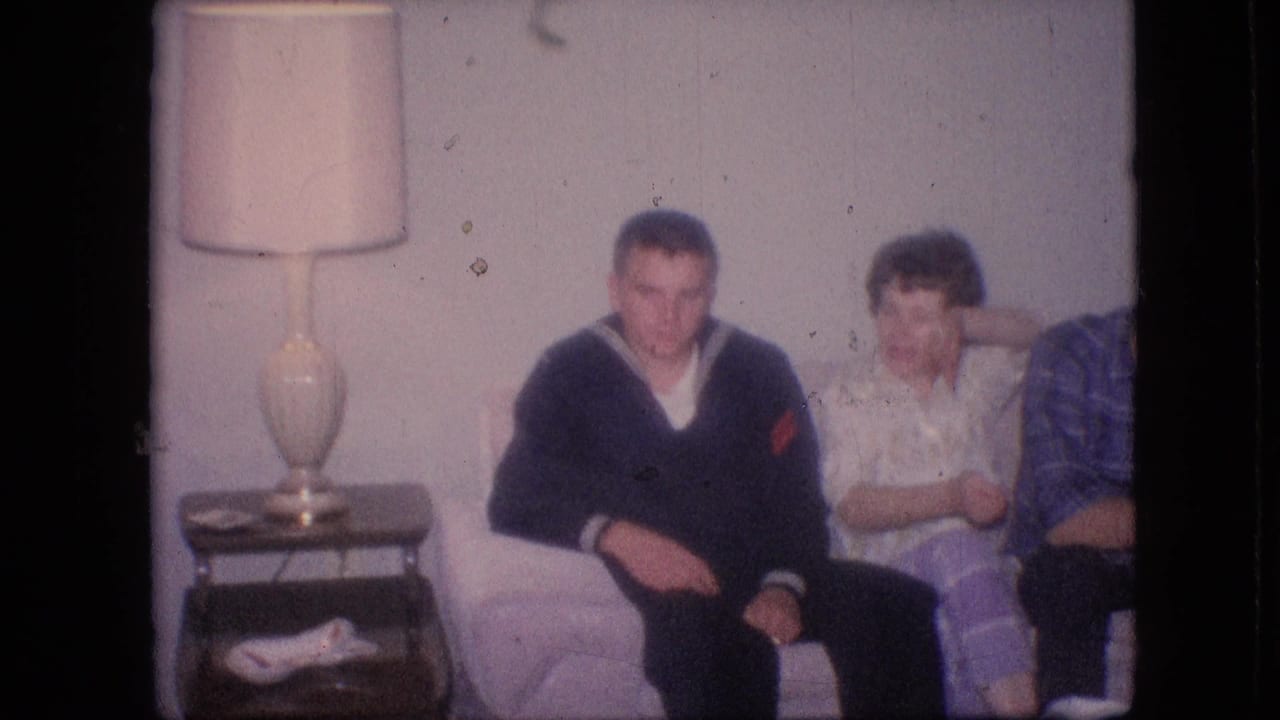 1964 Philadelphia home-movie frame showing adults seated on a living-room couch, with a man in a dark sweater beside a woman in light purple pants and another person partly visible at right.