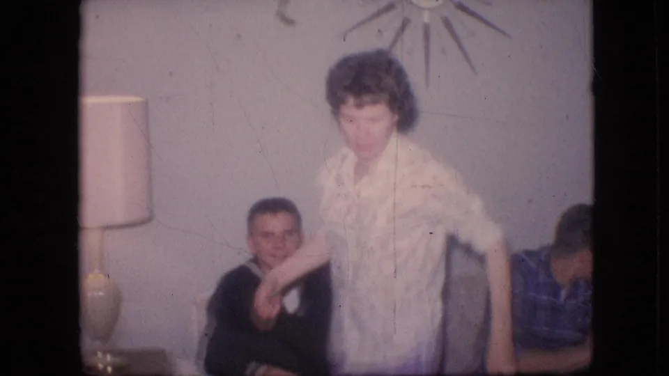 1964 Philadelphia home-movie frame showing a woman in a floral blouse and light purple pants moving across the living room, with a seated man, a partially visible person in plaid, and a starburst wall decoration behind her.