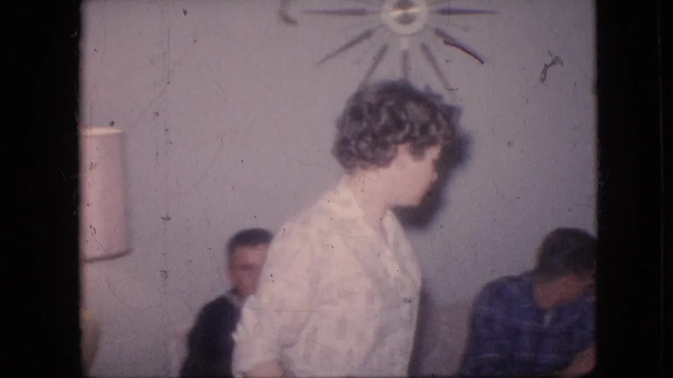 1964 Philadelphia home-movie frame showing the woman in a floral blouse passing close to the camera, partially obscuring seated adults on the couch beneath a starburst wall decoration.