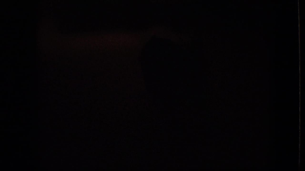An underexposed 8mm frame from the 1964 Philadelphia home movie, with the interior scene largely obscured in darkness and no clear subject detail visible.