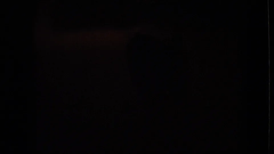 An underexposed 8mm frame from the 1964 Philadelphia home movie, with the interior scene largely obscured in darkness and no clear subject detail visible.
