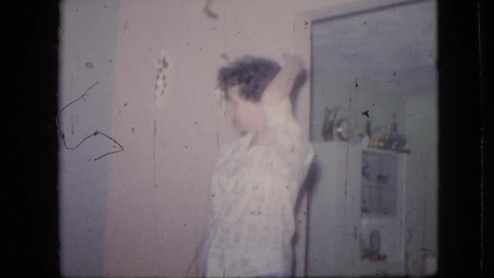 A person appears blurred in a Philadelphia living room from the 1964 8mm home movie, with a mirror, plant, and household shelf or cabinet visible behind them.
