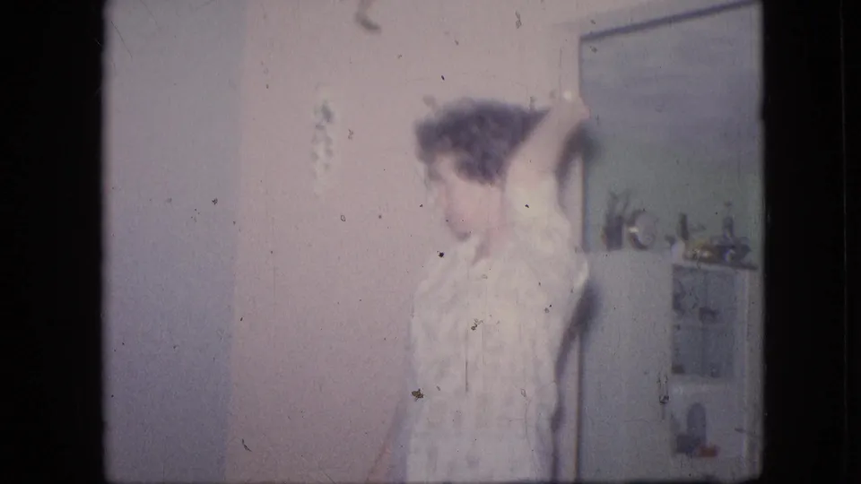 A blurred person stands or moves in the 1964 Philadelphia home interior, with a wall mirror, small plant, shelf or cabinet, and pale wall visible in the soft 8mm frame.