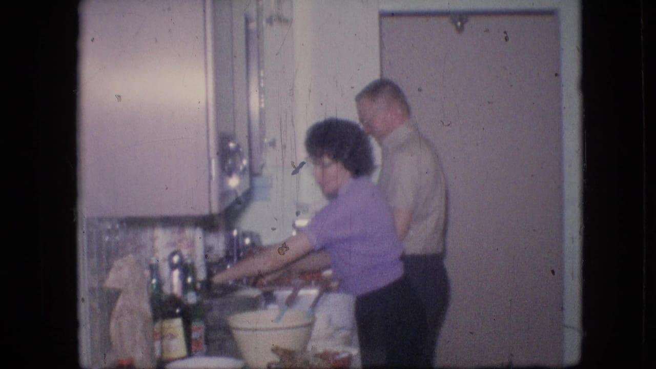 In a 1964 Philadelphia home-movie frame, a woman in a purple short-sleeved shirt works at the kitchen sink while a man in a light shirt stands behind her in a pastel private kitchen.