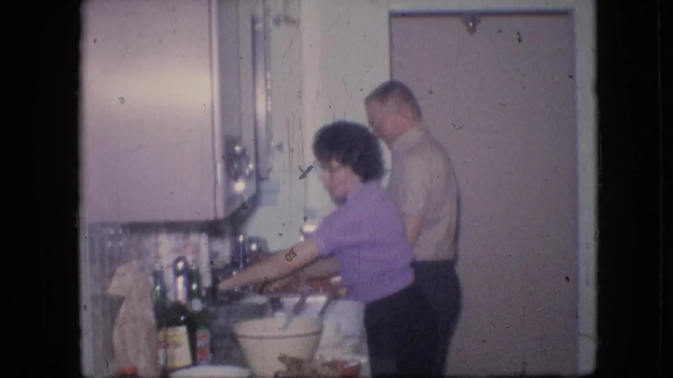 In a 1964 Philadelphia home-movie frame, a woman in a purple short-sleeved shirt works at the kitchen sink while a man in a light shirt stands behind her in a pastel private kitchen.