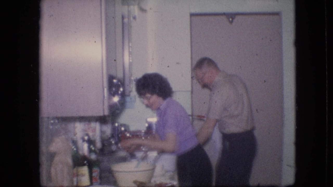 The frame shows the couple in the same 1964 Philadelphia kitchen, with the woman handling items at the sink and the man standing near the door behind her.