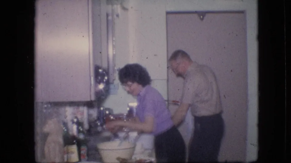 The frame shows the couple in the same 1964 Philadelphia kitchen, with the woman handling items at the sink and the man standing near the door behind her.