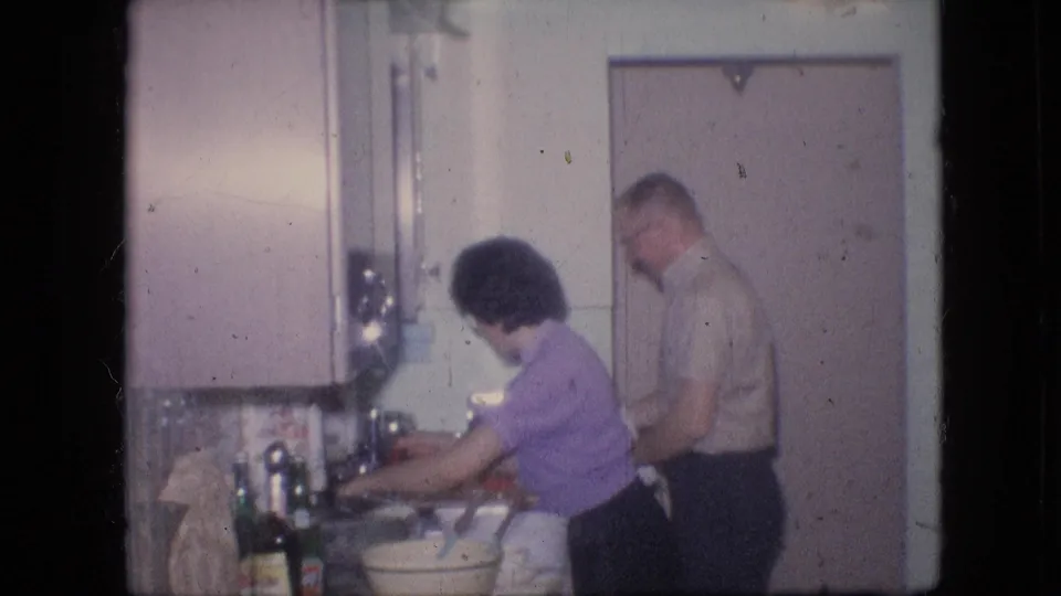 A woman leans toward the sink in a pastel kitchen while a man stands behind her, with cabinets, bottles, a bowl, and towels visible in the 8mm home-movie frame.